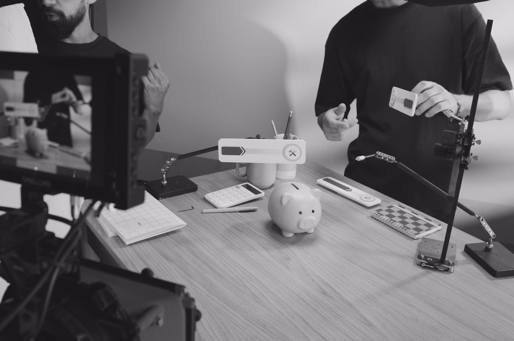 Two people at a desk with a piggy bank, microphone, and camera during a podcast or video recording session