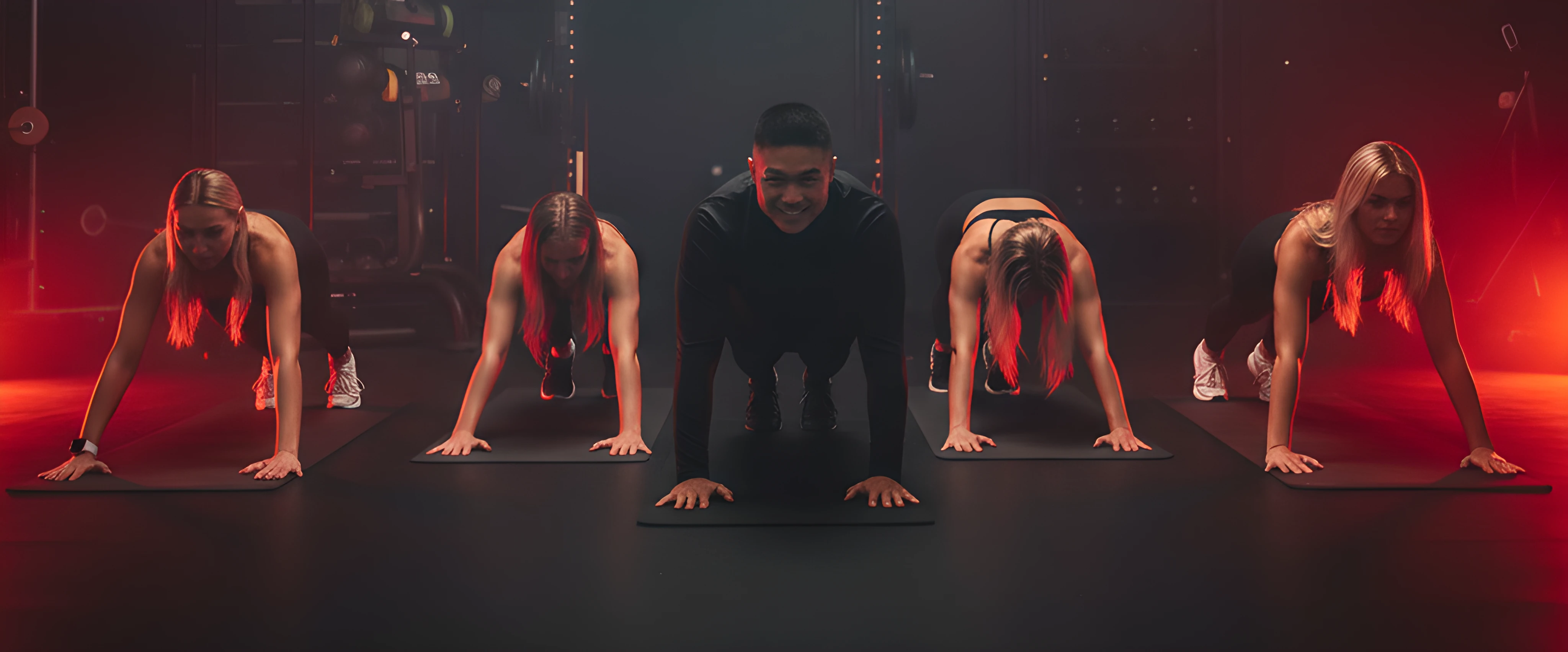 Five people in a gym performing plank exercises on black mats under red lighting.