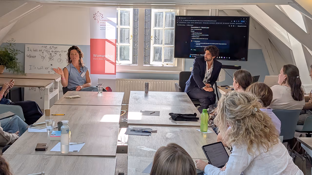 Groep mensen in een lichte vergaderruimte met een vrouw die spreekt en een man die luistert naast een scherm met tekst.