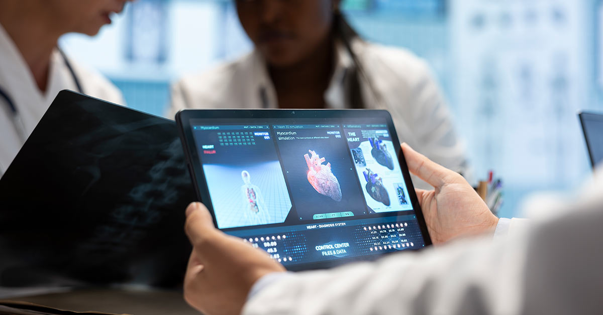 Médico segurando um tablet com imagens 3D de um coração e dados clínicos na tela, enquanto outros profissionais de saúde conversam ao fundo em um ambiente hospitalar.