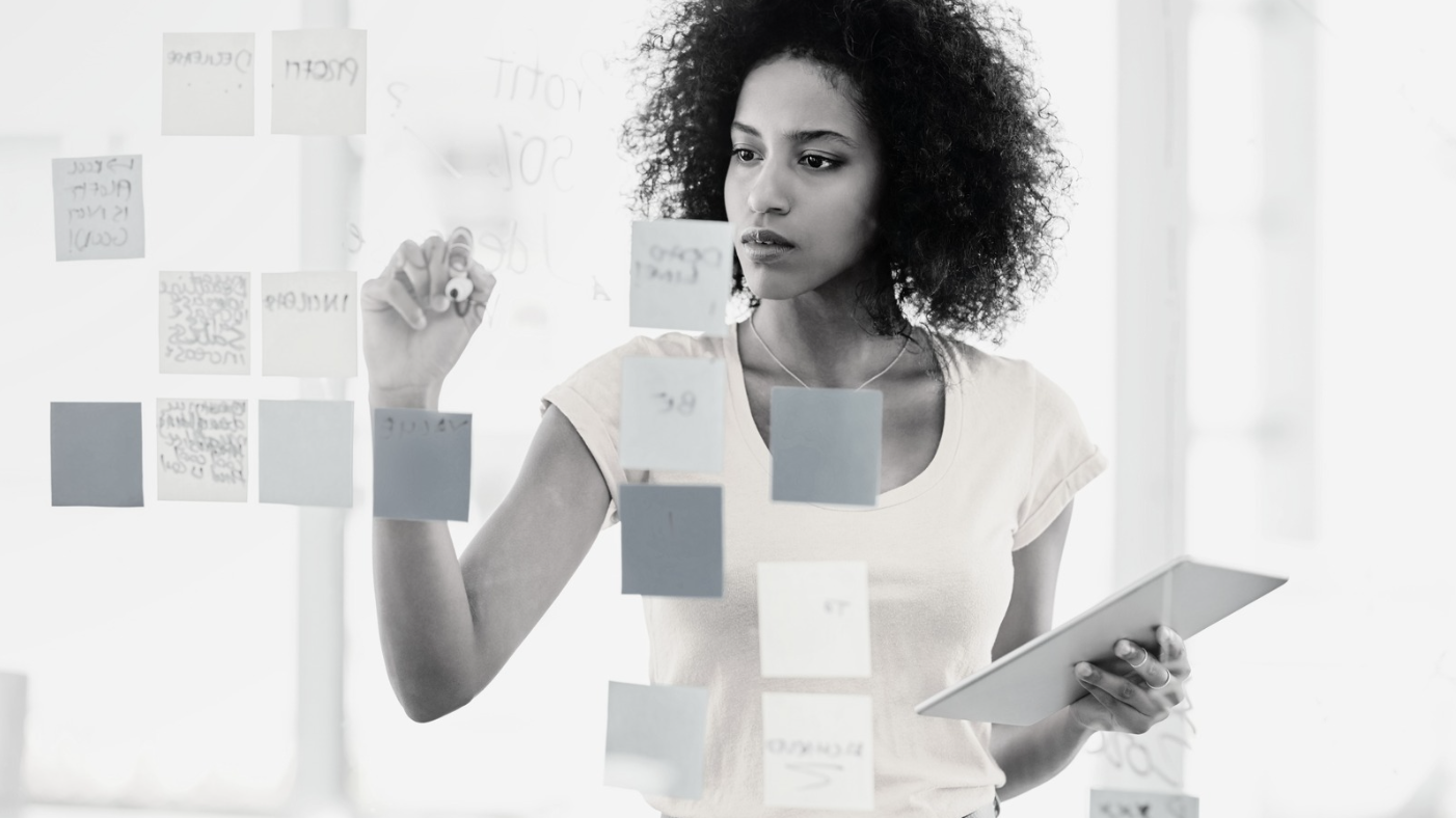 Focused woman holding a tablet and organizing sticky notes on a glass wall, symbolizing planning and creative problem-solving in a bright workspace.