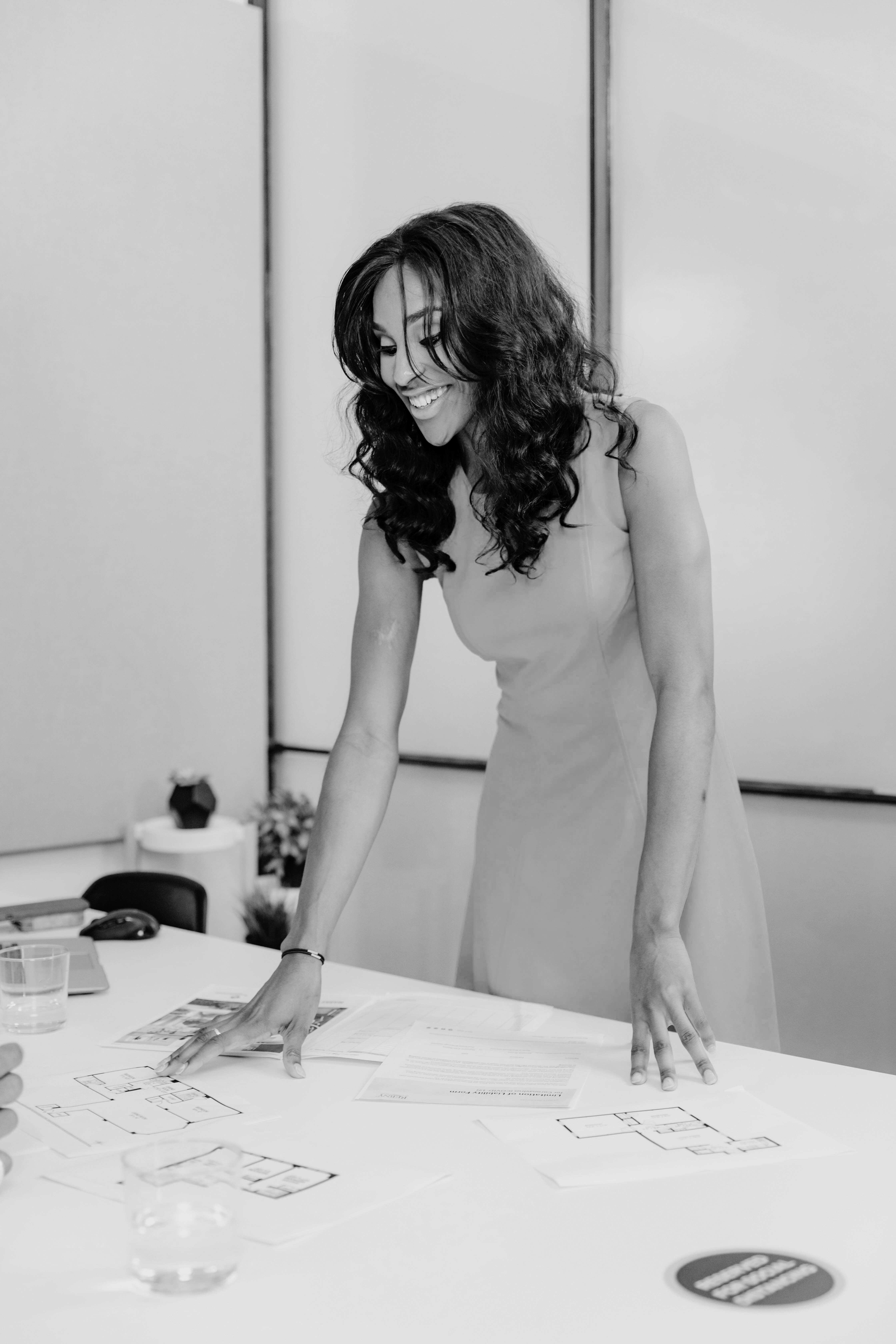 Black-and-white image of a smiling woman standing at a table, reviewing design layouts and documents, exuding confidence and professionalism in a collaborative workspace.