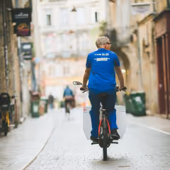 Homme en polo bleu livrant un colis en faisant du vélo dans une rue pavée en ville.