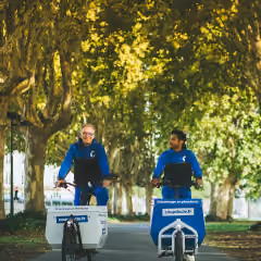 Deux hommes à vélo cargo se déplaçant sous des arbres aux feuilles vertes.