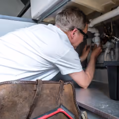 Un plombier réparant des tuyaux sous un évier avec une trousse d’outils à proximité.