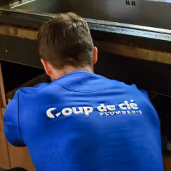 Homme portant un t-shirt bleu avec le logo Coup de clé Premiers au dos, accroupi devant un évier.