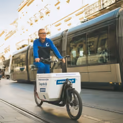 Personne portant un sweat bleu et des lunettes conduisant un vélo-cargo électrique blanc dans une rue urbaine avec un tramway en arrière-plan.