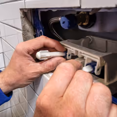 Deux mains réparent ou installent un WC encastré dans un mur carrelé blanc.