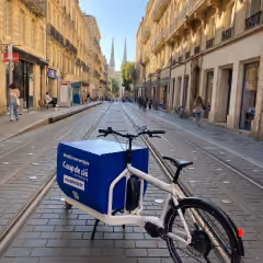 Vélo cargo blanc garé sur une rue pavée avec des immeubles de chaque côté et une église à deux flèches au loin.