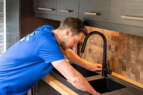 Un homme en t-shirt bleu répare ou inspecte un évier de cuisine noir avec un robinet moderne noir.