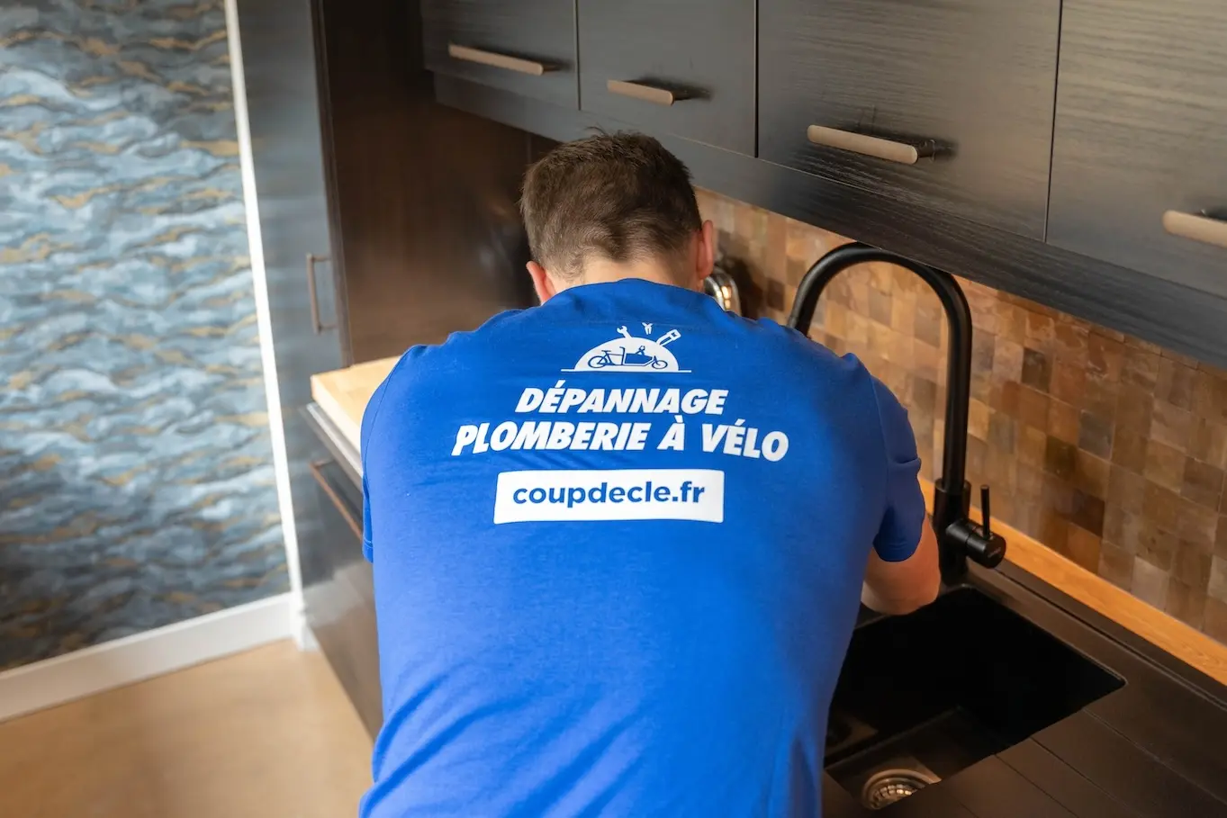 Plombier en t-shirt bleu avec un marquage coup de clé dans le dos intervenant sur un évier de cuisine