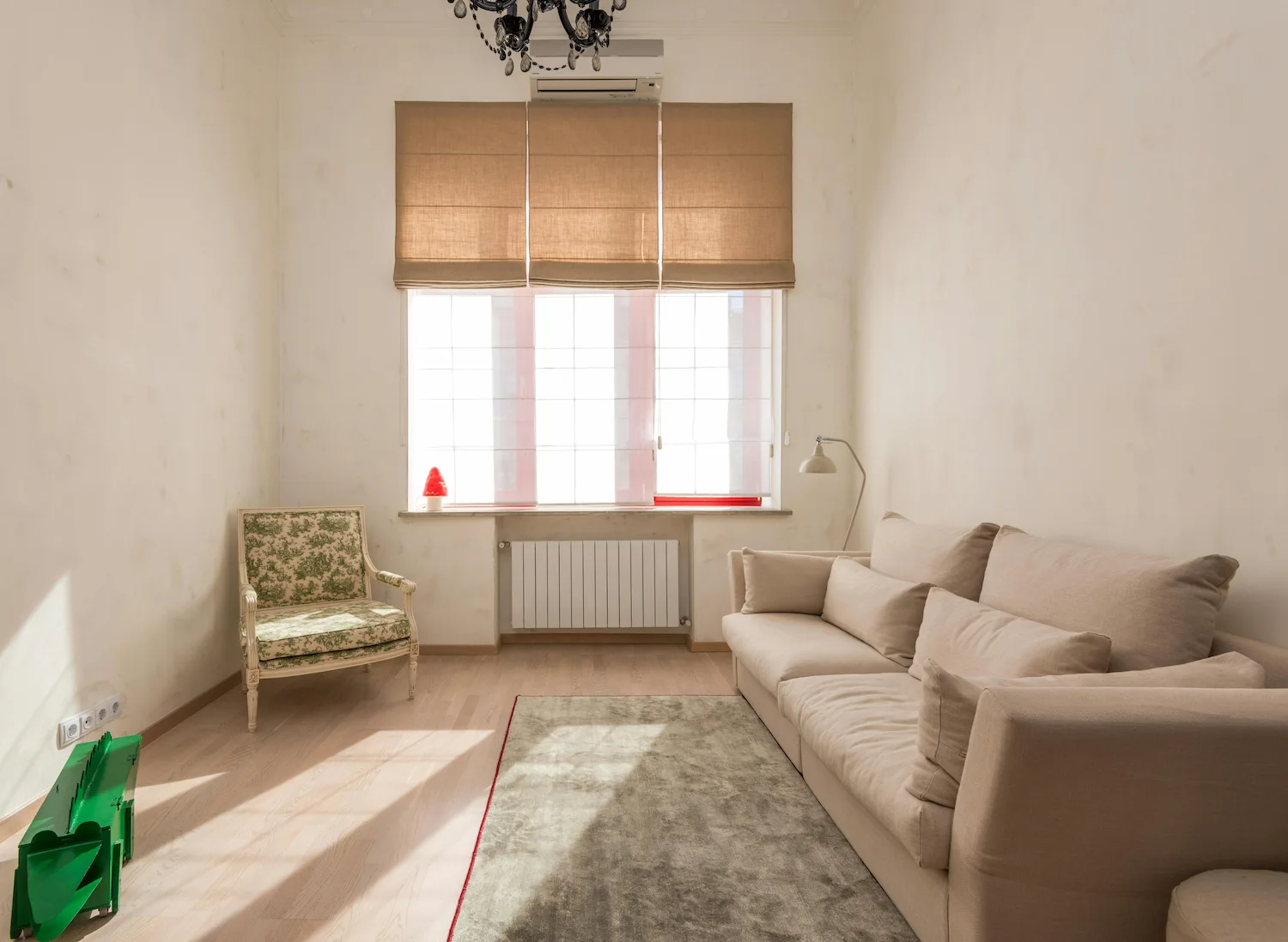 Salon lumineux avec un canapé beige, un fauteuil vert à motifs, un tapis gris, un radiateur sous la fenêtre et un lustre noir.