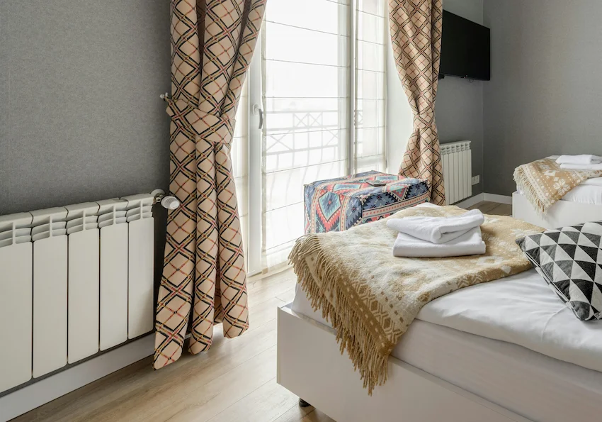 Chambre avec deux lits faits, couvertures beiges à motifs et serviettes blanches pliées, rideaux à carreaux, radiateurs blancs et télévision murale.