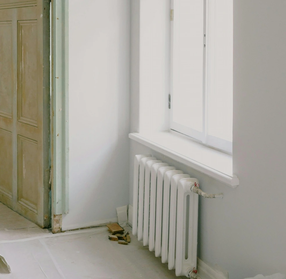 Radiateur blanc à colonnes installé sous une fenêtre dans une pièce en cours de rénovation avec sol protégé et portes anciennes.