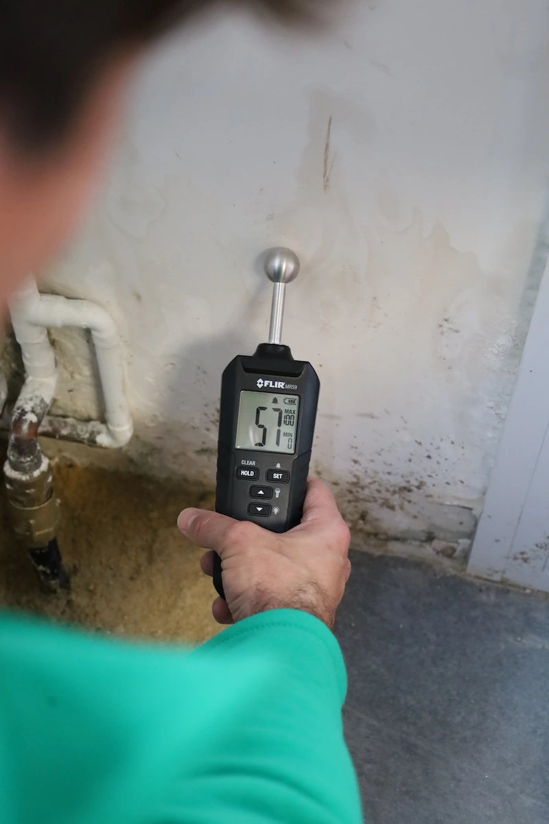 Une main tenant un hygromètre numérique FLIR MR59 affichant une humidité de 57 % devant un mur avec des traces d'humidité.