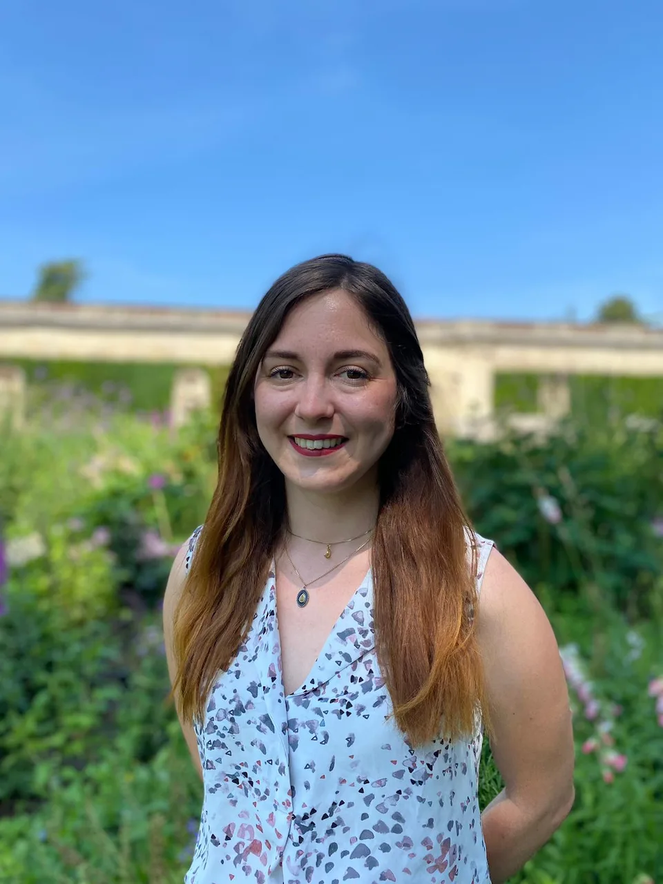 Jeune femme souriante aux cheveux longs portant un débardeur blanc à motifs, posant dans un jardin fleuri sous un ciel bleu clair.