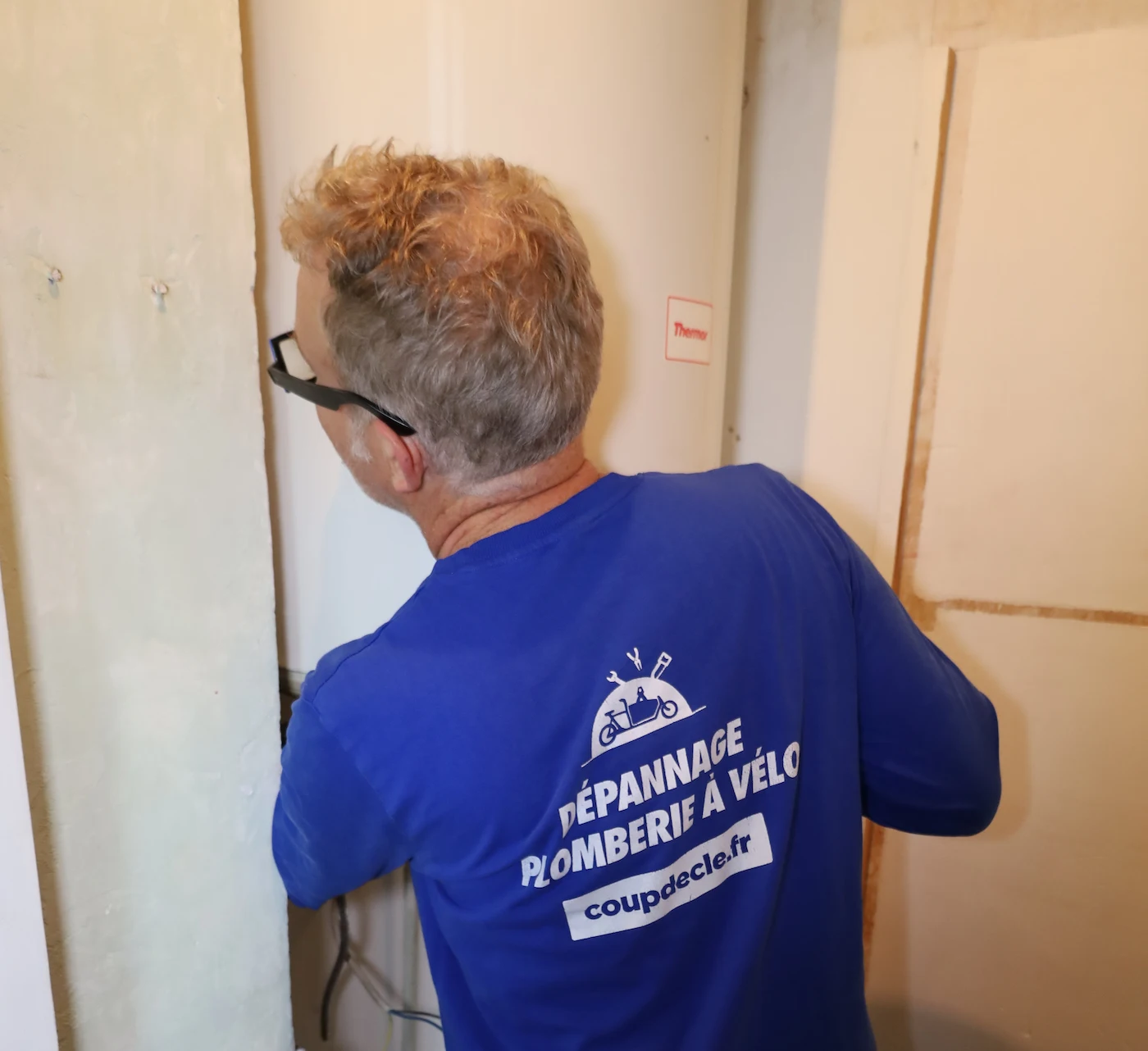 Un plombier portant un t-shirt bleu avec le texte « Dépannage plomberie à vélo coupdecle.fr » travaillant sur un chauffe-eau mural.
