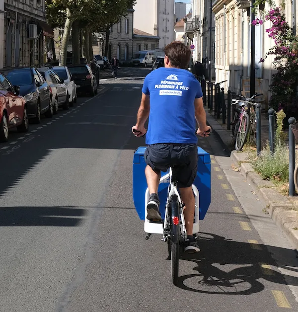 Homme en t-shirt bleu faisant de la plomberie à vélo dans une rue urbaine.