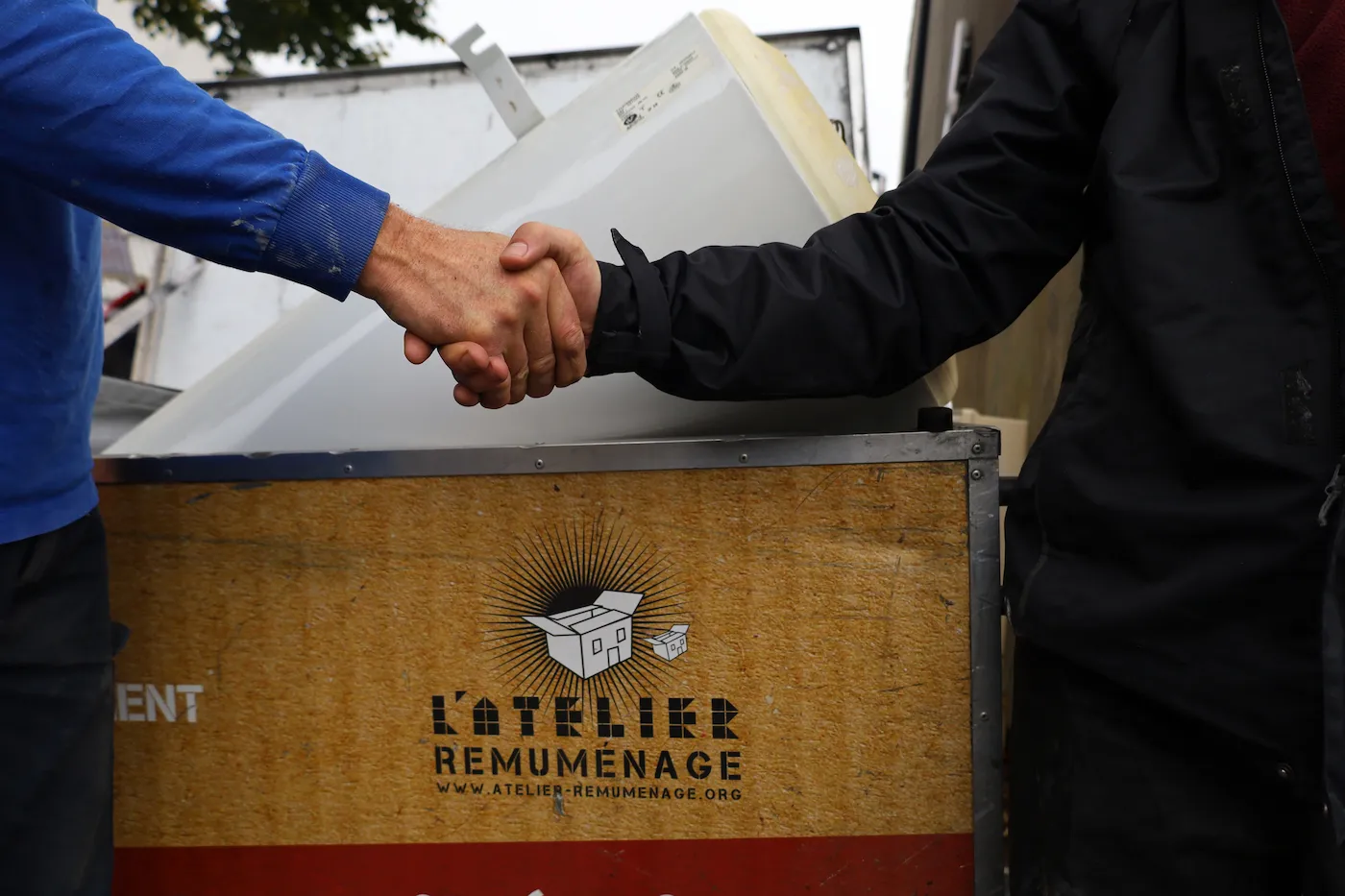 Deux personnes se serrant la main devant un panneau en bois avec le logo et le texte de l'atelier Remuménage.