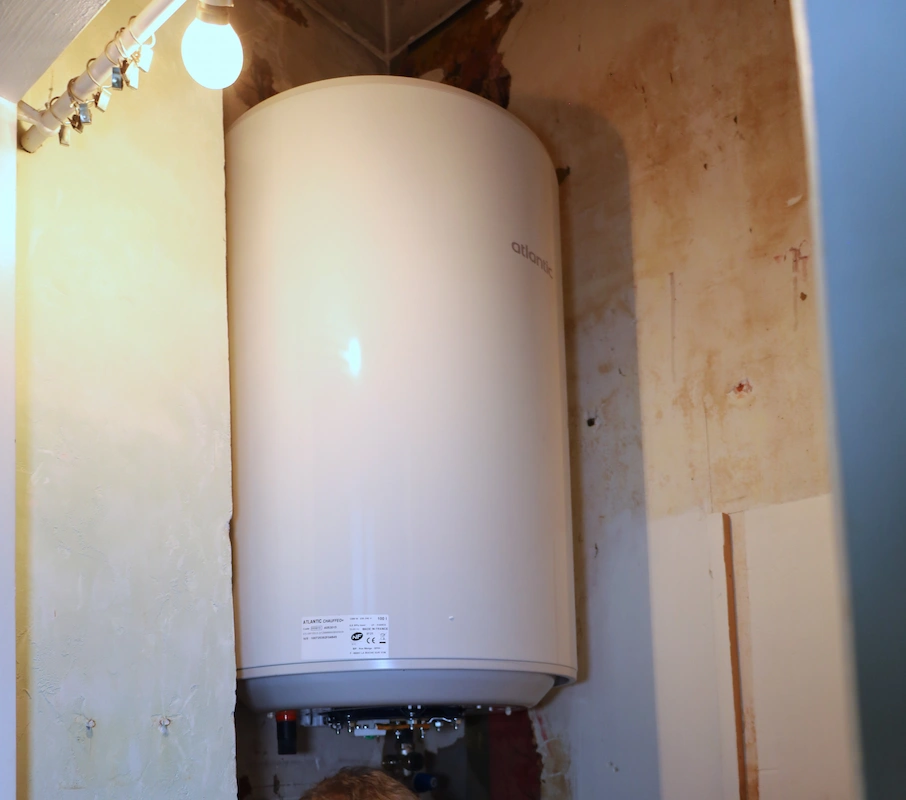 Chauffe-eau mural blanc installé dans un coin d'une pièce avec mur en plâtre et lumière suspendue.