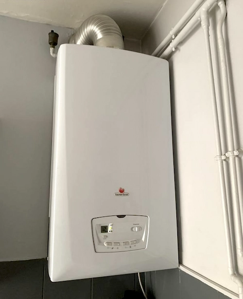 Chaudière à gaz blanche montée sur un mur avec tuyau d'évacuation en métal ondulé en haut et des tuyaux blancs à droite.