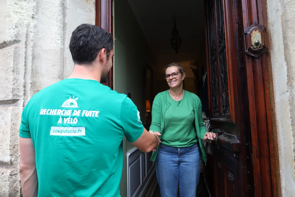 Un technicien en t-shirt vert avec l'inscription « Recherche de fuite à vélo » serre la main d'une femme souriante à la porte d'entrée d'une maison.