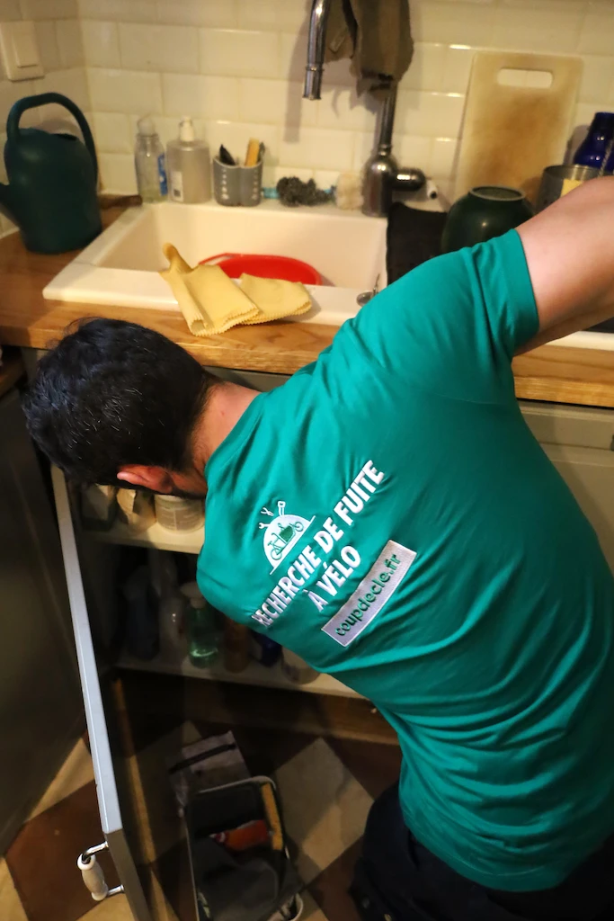 Technicien en polo vert cherchant une fuite sous un évier de cuisine avec des produits de nettoyage visibles sur le plan de travail.