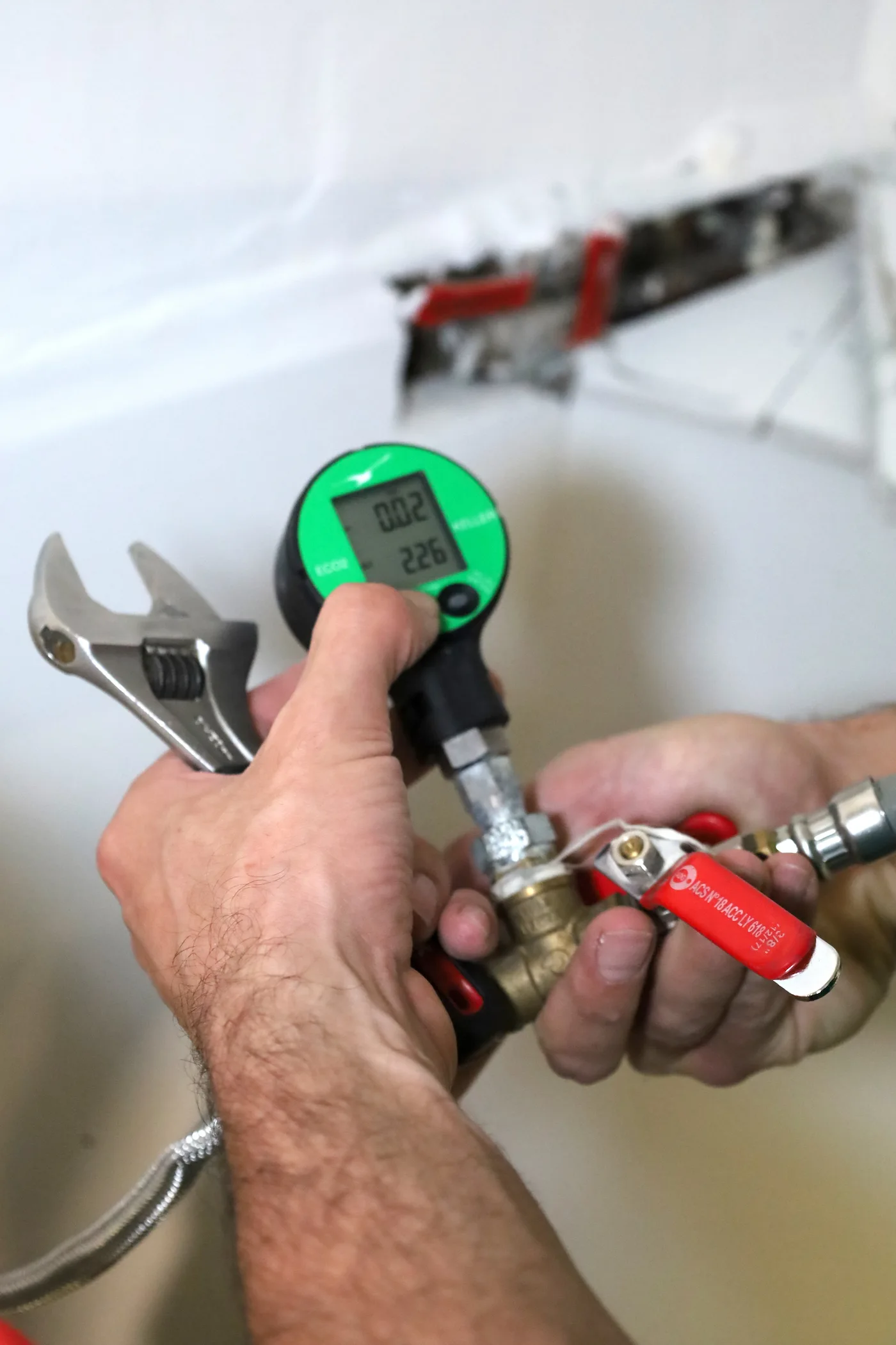 Main tenant un clé anglaise et un manomètre numérique relié à une vanne avec une poignée rouge lors d'un test de pression.