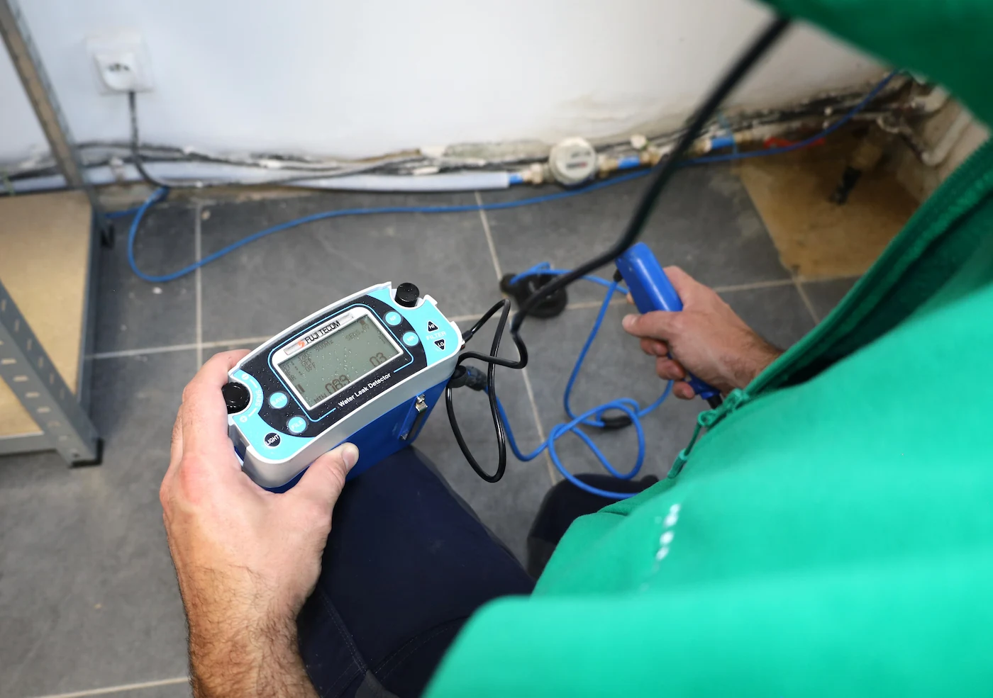 Technicien utilisant un détecteur de fuites d'eau électro-acoustique avec un écran et une sonde dans une pièce carrelée.
