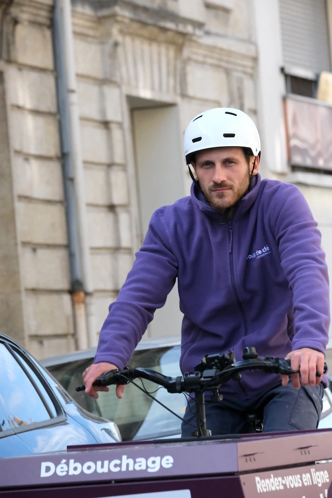 Homme portant un casque blanc et un sweat violet, tenant le guidon d'un vélo avec un panneau indiquant 'Débouchage' sur un fond urbain.