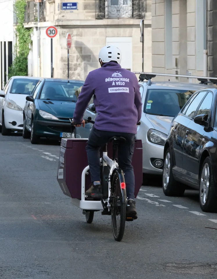 Technicien portant un casque blanc en vélo cargo violet, vu de dos, circulant dans une rue urbaine avec des voitures garées.