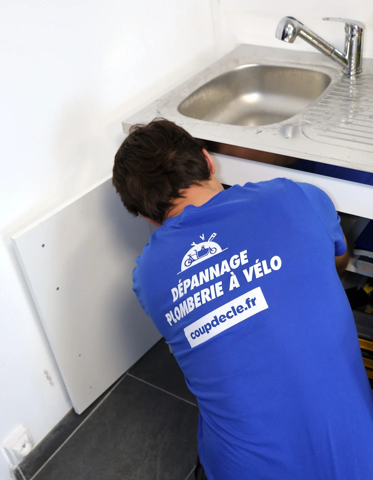 Plombier en polo bleu réparant une plomberie sous un évier de cuisine.