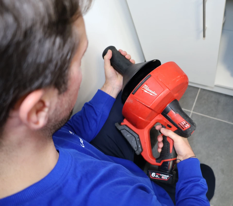 Un homme en sweat bleu tient un déboucheur électrique rouge de la marque Milwaukee dans une pièce avec un sol carrelé gris.