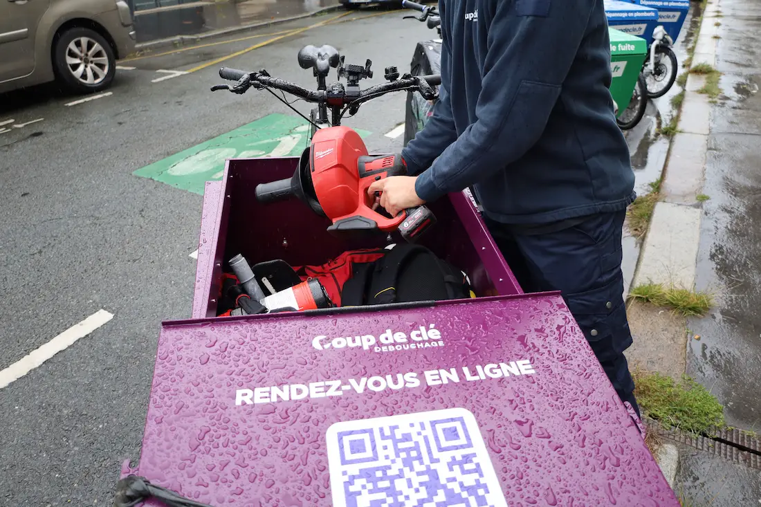 Personne tenant une clé à choc rouge dans la caisse avant d'un vélo cargo violet avec la mention « Coup de clé Débouchage RENDEZ-VOUS EN LIGNE » et un QR code.