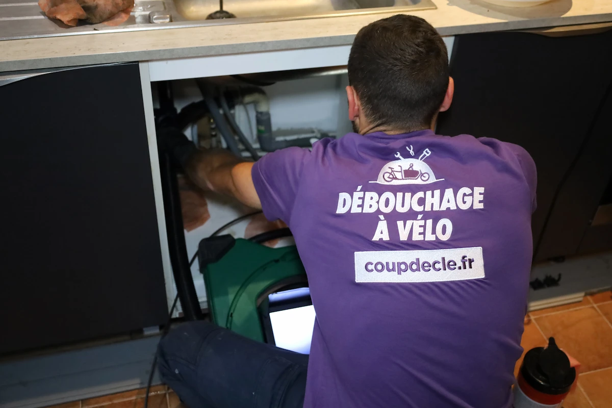 Technicien portant un t-shirt violet avec l'inscription 'Débouchage à vélo' travaillant sous un évier de cuisine.