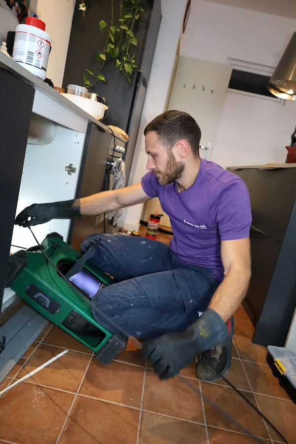 Un plombier en t-shirt violet et gants noirs répare une canalisation sous l'évier dans une cuisine moderne.