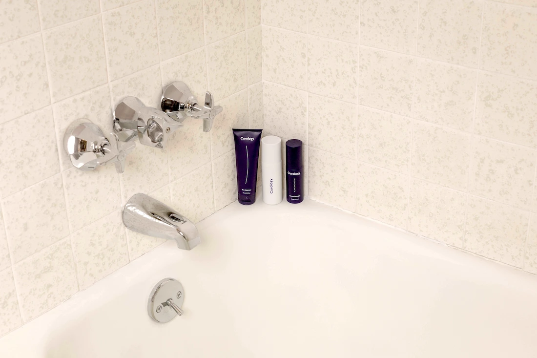 Trois produits Curology posés dans le coin d'une baignoire blanche avec robinetterie chromée et carrelage beige.