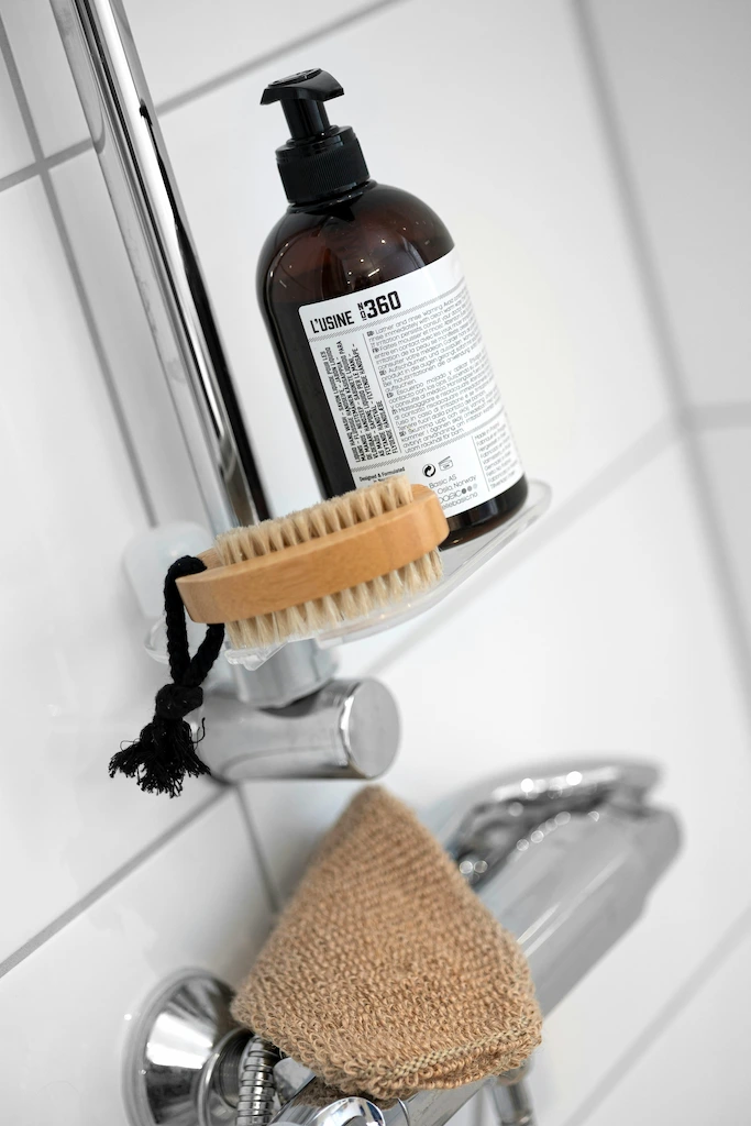 Flacon pompe de savon L'USINE 360 et brosse en bois suspendus près d'un robinet de douche chromé avec un gant de toilette beige.