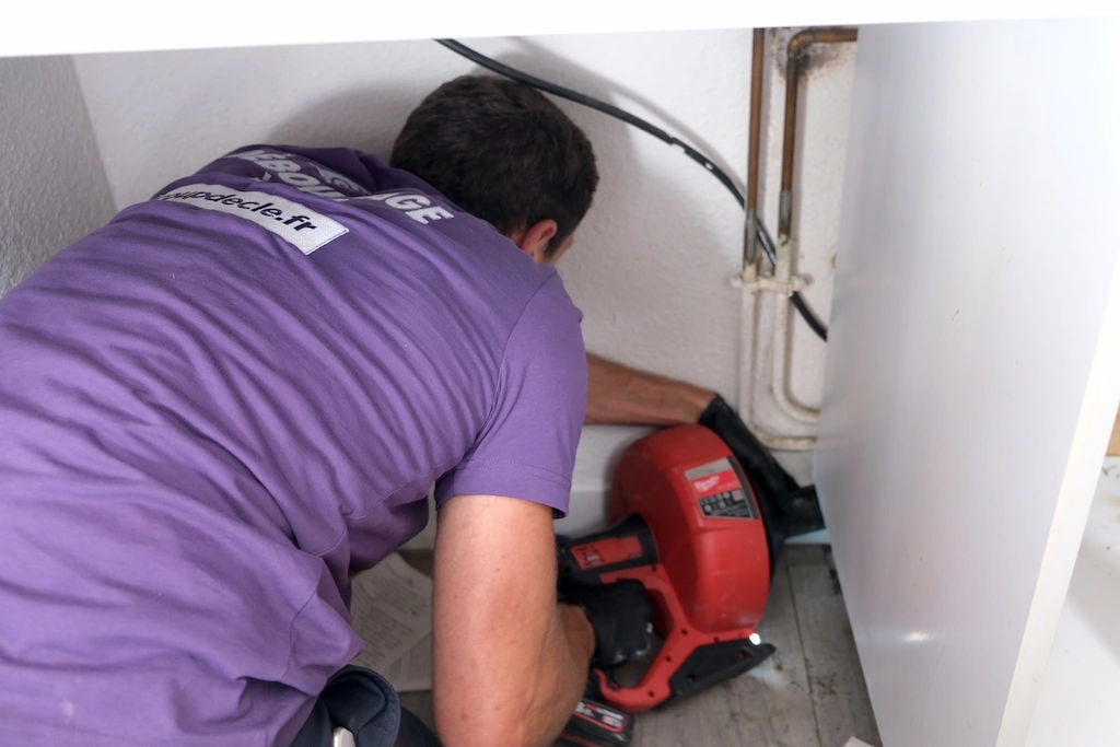 Un plombier en t-shirt violet utilise un outil électrique rouge pour déboucher une canalisation sous un évier.