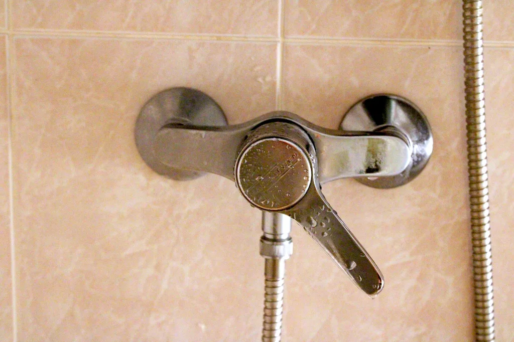 Robinet mitigeur de douche en métal fixé sur un mur carrelé beige avec un flexible métallique.