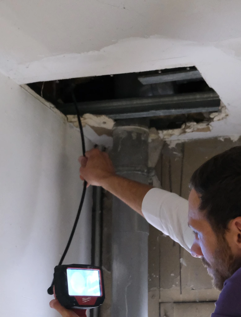 Un homme utilise une caméra d'inspection pour examiner un tuyau d'évacuation visible à travers une ouverture dans le plafond.