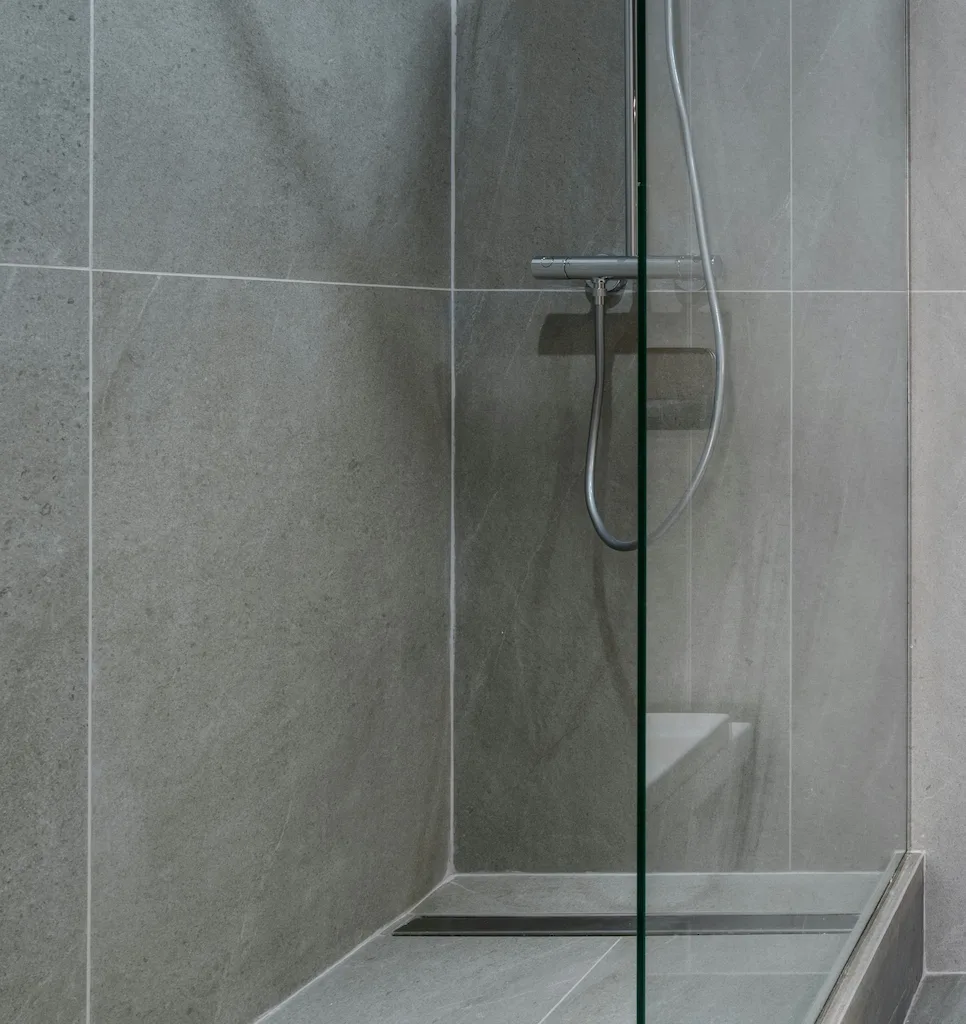 Douche moderne avec murs et sol en carrelage gris, paroi de verre et pomme de douche en métal.