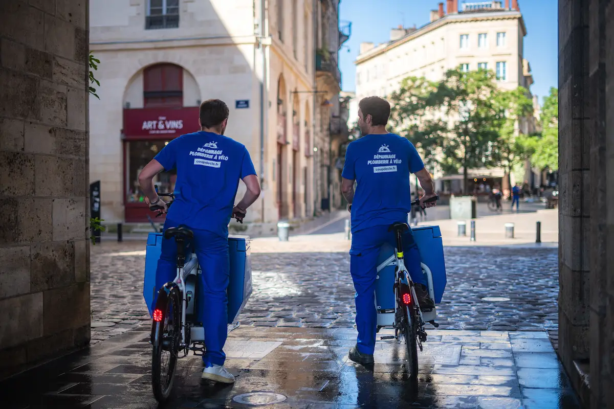 Deux techniciens en tenue bleue sur des vélos avec des caisses bleues dans une rue pavée d'une ville.