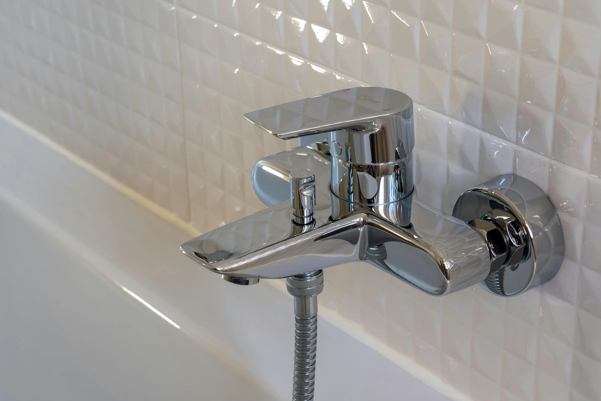 Robinet mitigeur en chrome fixé au mur au-dessus d'une baignoire blanche avec carrelage blanc à motifs géométriques en arrière-plan.