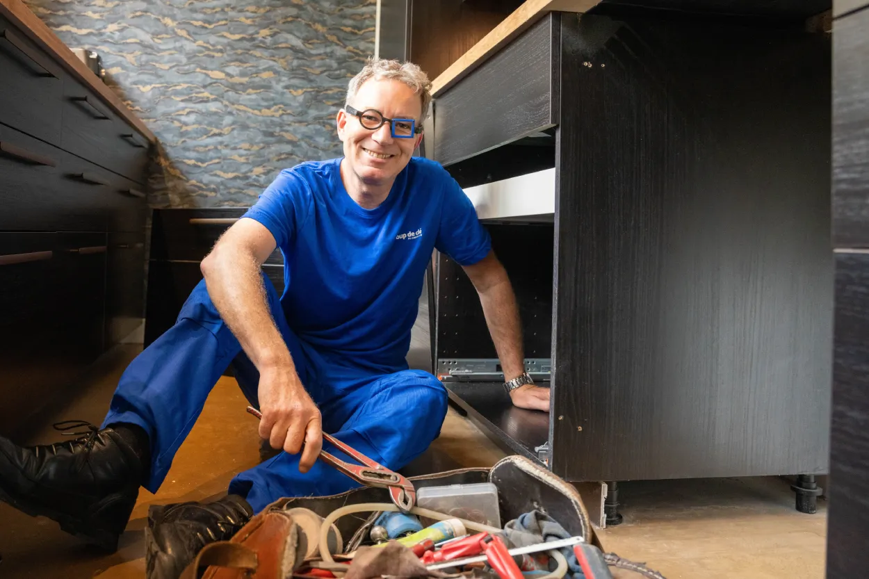 Plombier souriant en bleu, accroupi devant un meuble noir ouvert avec une clé anglaise en main.