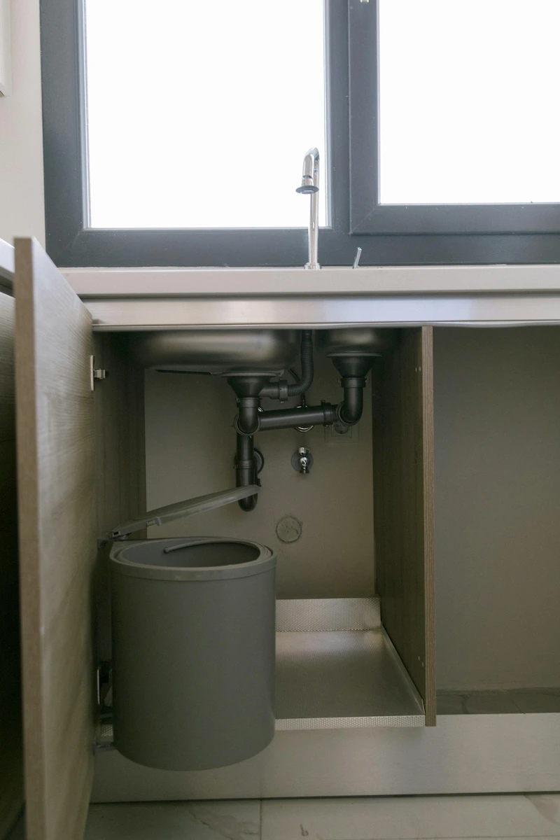 Canalisations sous un évier avec poubelle intégrée à la porte du meuble ouverte.