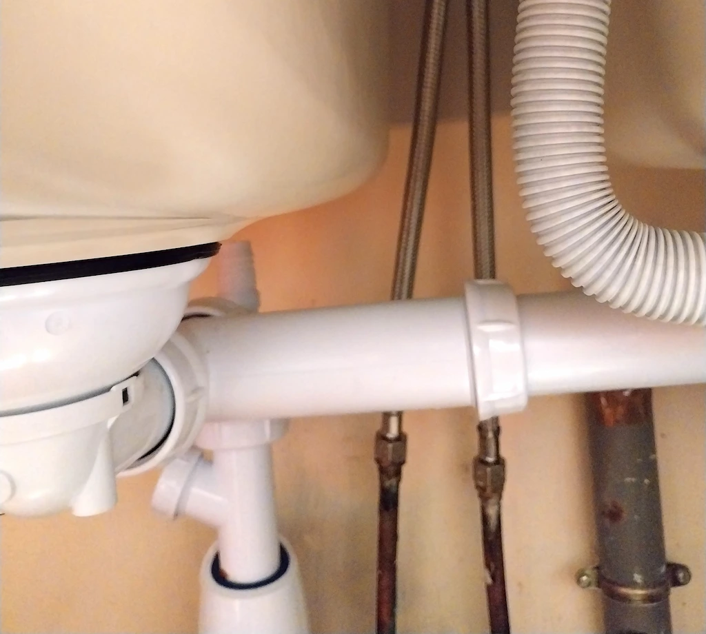 Vue rapprochée des tuyaux de plomberie sous un évier en plastique blanc avec un tuyau flexible ondulé.