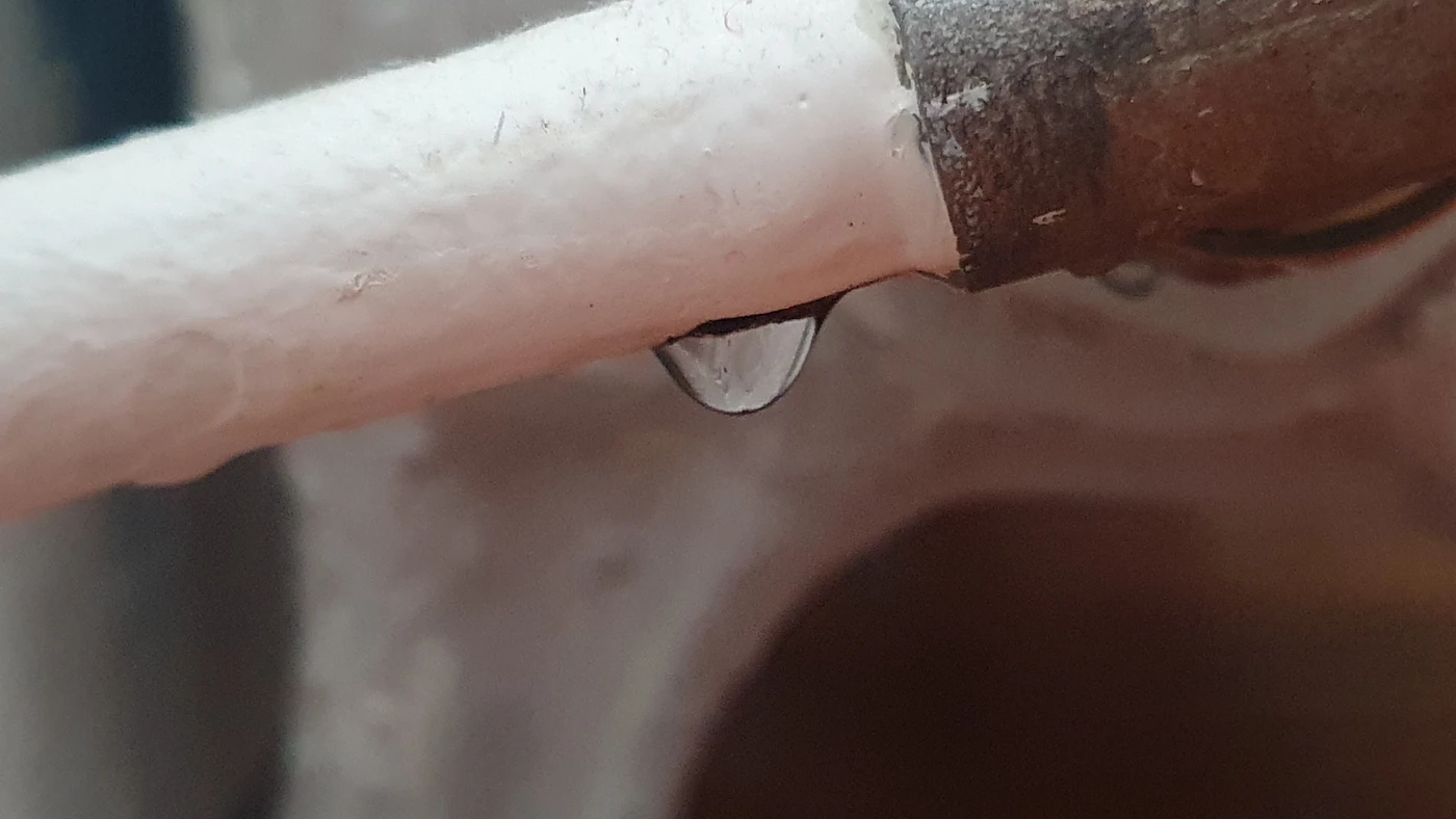Goutte d'eau suspendue à une fuite entre un tuyau blanc et une connexion métallique rouillée.