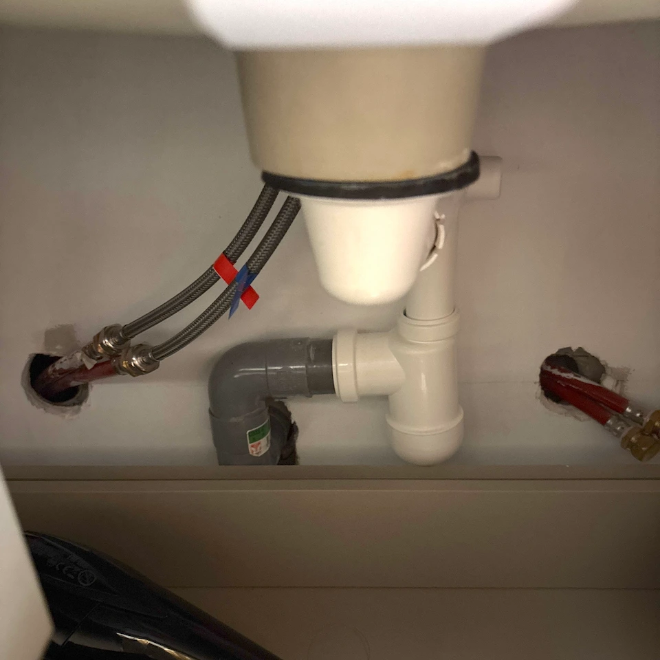 Sous un évier, tuyaux d’alimentation flexibles reliés à un broyeur d’évier et tuyau de sortie en PVC gris connecté au système de plomberie.