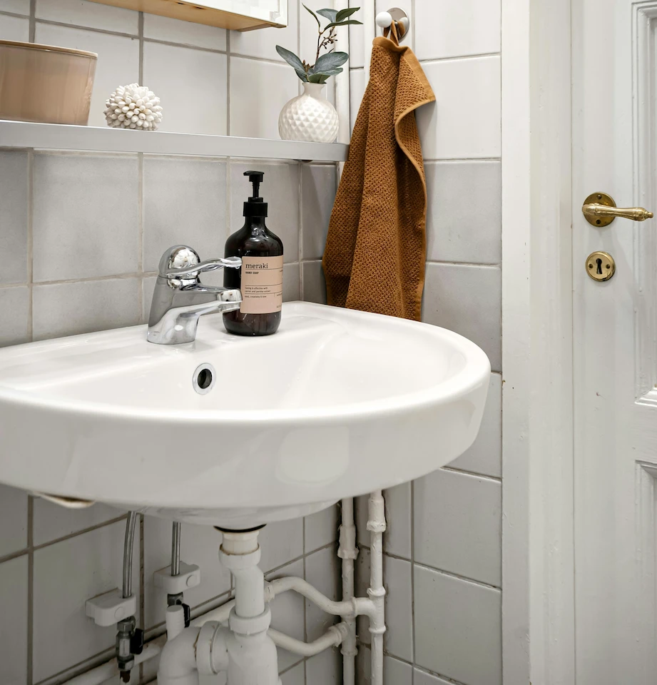 Lavabo blanc fixé au mur dans une salle de bain avec robinet chromé, bouteille de savon liquide foncée, serviette marron suspendue, carreaux gris clair, et porte blanche avec poignée dorée.